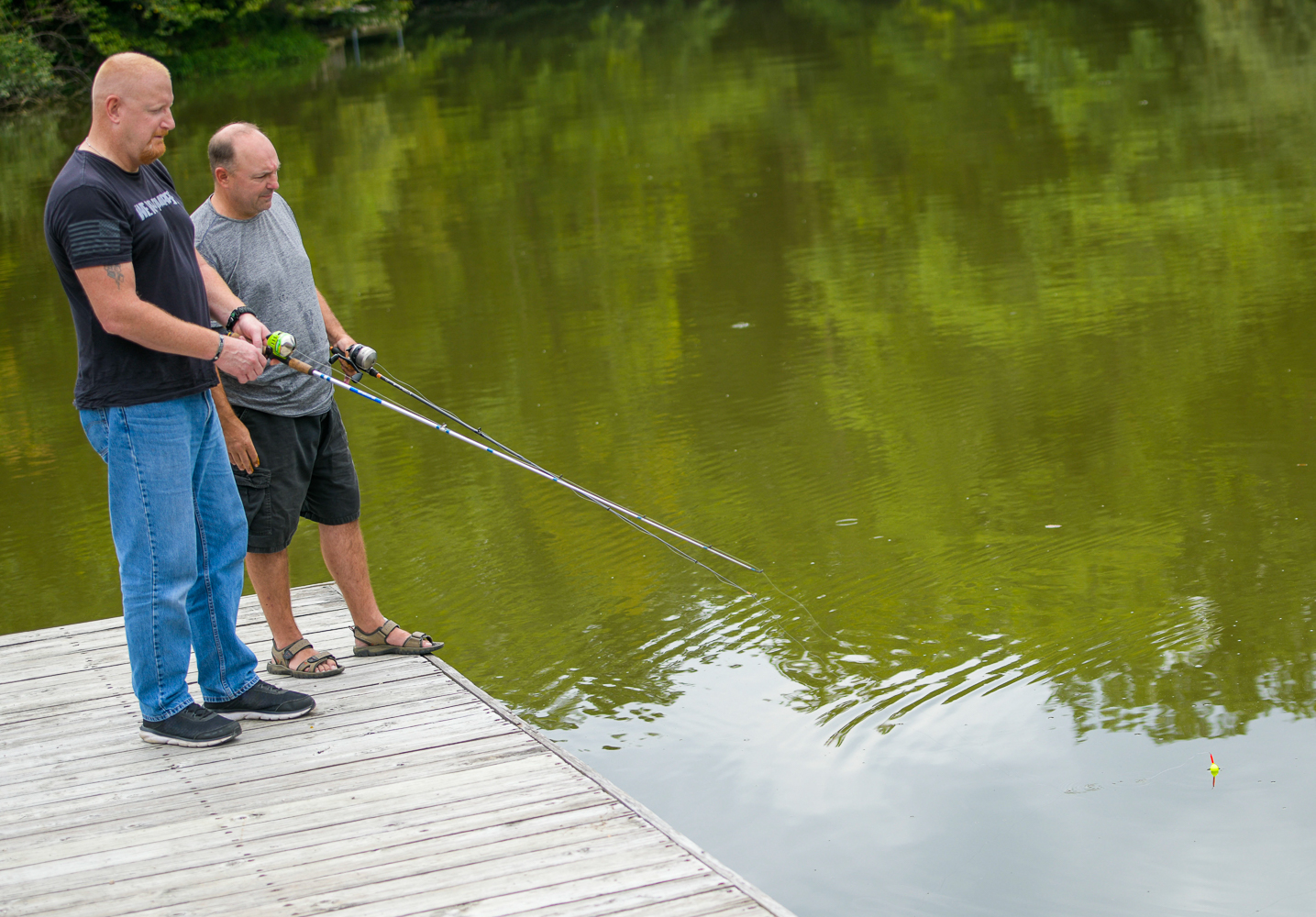 Two white men stand together fishing in a lake. The taller one is wearing sneakers, blue jeans and a black shirt. The shorter one is wearing sandals, black shorts and a grey t-shirt.