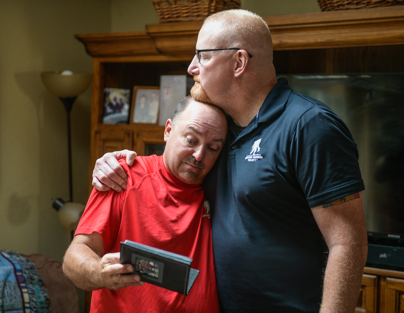 Two white men stand embracing. The taller one on the right has his chin on the shorter man's head, and his arm around his shoulder. The shorter man is holding a photo album. The shorter man on the left is wearing a red t-shirt and the taller man on the right is wearing a black polo short sleeve shirt. Both shirts have the logo of Wounded Warrior Project on them.