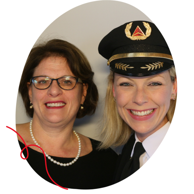 A white woman with short brown hair wearing a black top and a pearl necklace standing next to a woman with short blonde hair wearing a pilot uniform.