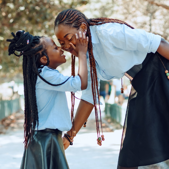 A Black child wearing a blue long sleeve button down wearing braids down her back and half of it as a bun up and a Black woman in a blue long sleeve button down and long braids.